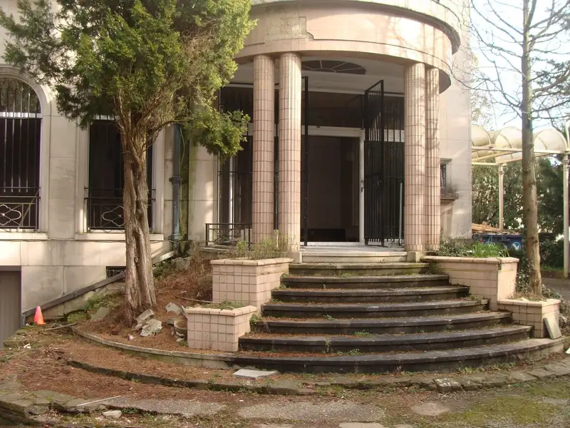 Portique d’entrée arrondi à quatre colonnes d’un bâtiment inoccupé ; herbes entre les marches, mousse sur le perron et un conteneur près de l’entrée.
