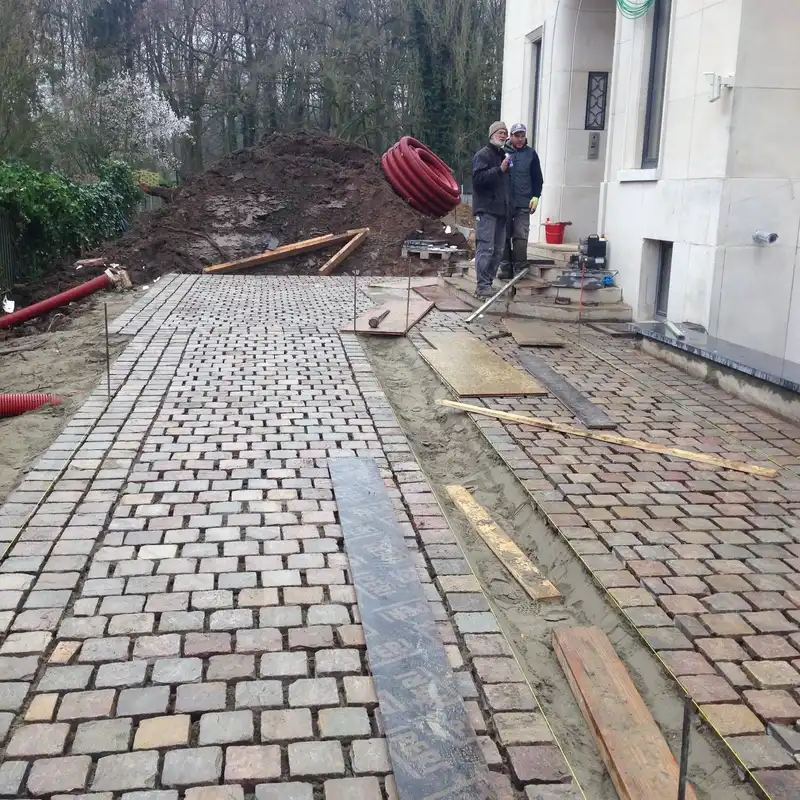 Cour avant en chantier : pavés posés en rangées sur lit de sable, cordeaux tendus et bandes sombres délimitant les surfaces.