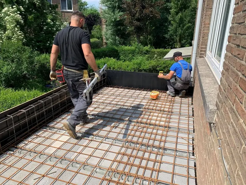 Deux membres de l’équipe posent le treillis d’armature sur la dalle de terrasse le long de la façade, jardin à l’arrière.