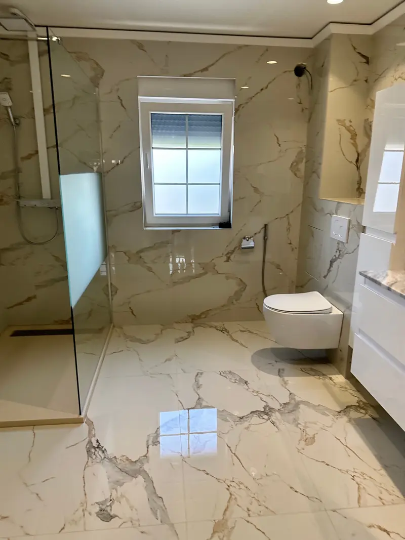 Salle de bains entièrement en marbre, douche à l’italienne derrière une paroi vitrée et fenêtre à volet.