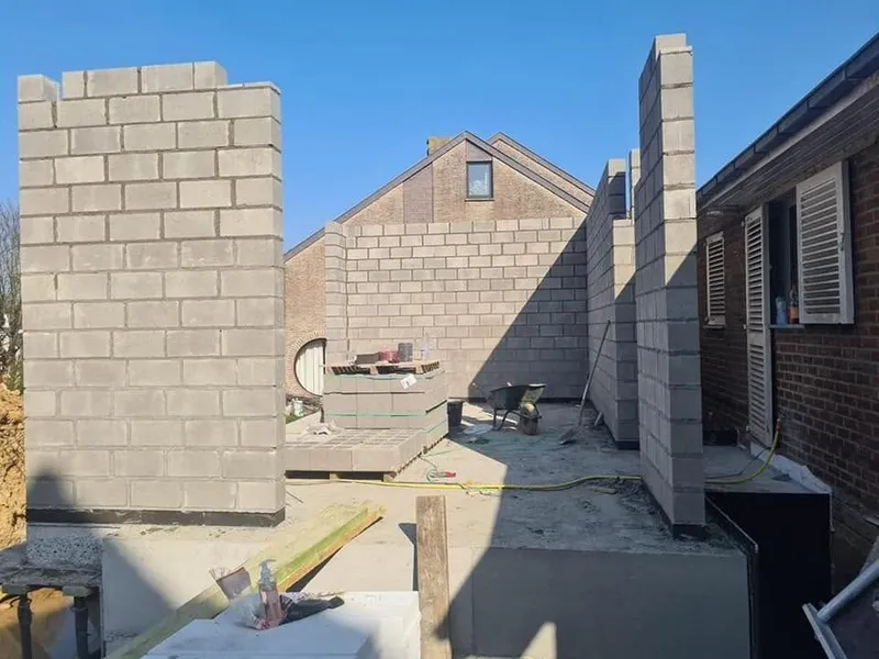 Maçonnerie en blocs d’une extension qui monte sur un ciel bleu profond, façade existante à côté.