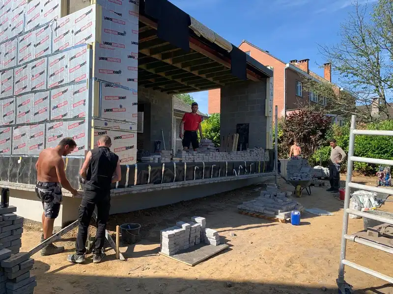Cinq membres de l’équipe devant l’extension en gros œuvre : la maçonnerie en blocs et le solivage portent à faux, palettes de brique grise prêtes sur le sable.